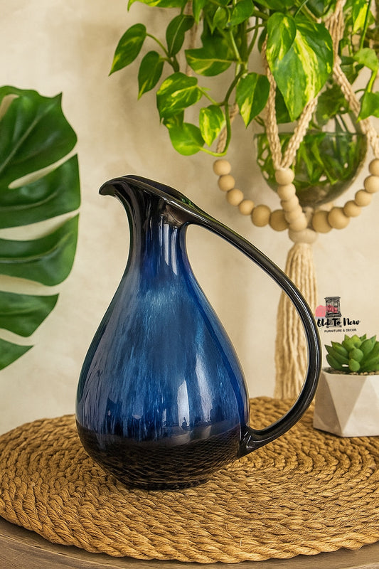 A deep blue ceramic pitcher with a sculptural curved handle, displayed in a boho-style setting with greenery and natural textures, perfect as a decorative vase or home accent.