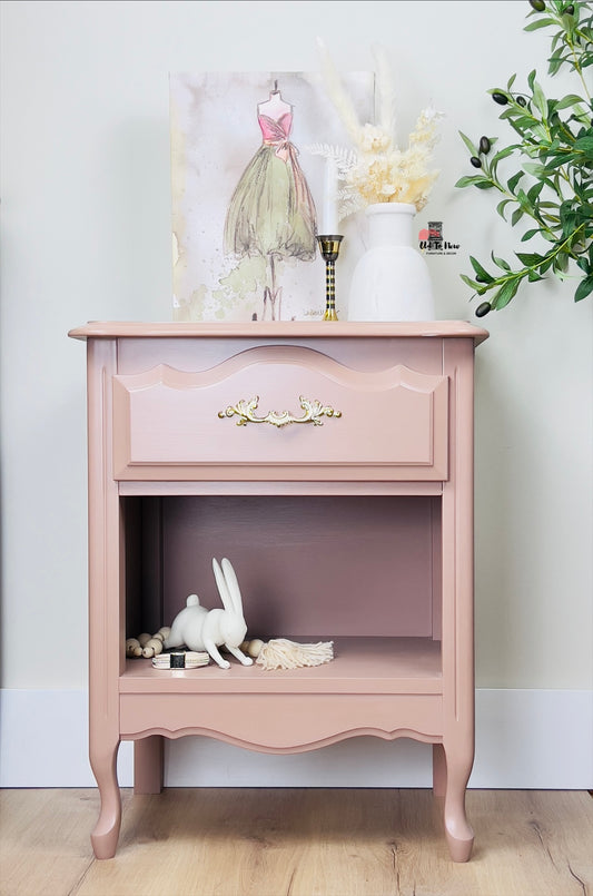 Blush pink vintage nightstand with curved legs, ornate brass drawer handle, and open bottom shelf, styled with a ceramic bunny and feminine artwork in a modern cottage setting; Old to New Furniture & Decor