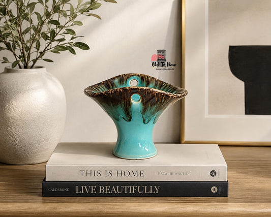 A turquoise and brown glazed ceramic vase holder with two circular cutouts, displayed on a stack of books on a wooden table next to a white textured vase with greenery.