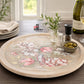 A round wooden serving tray with a floral and bird illustration, styled on a dark wood table with a wine bottle, glasses, olives, and a small vase of greenery.