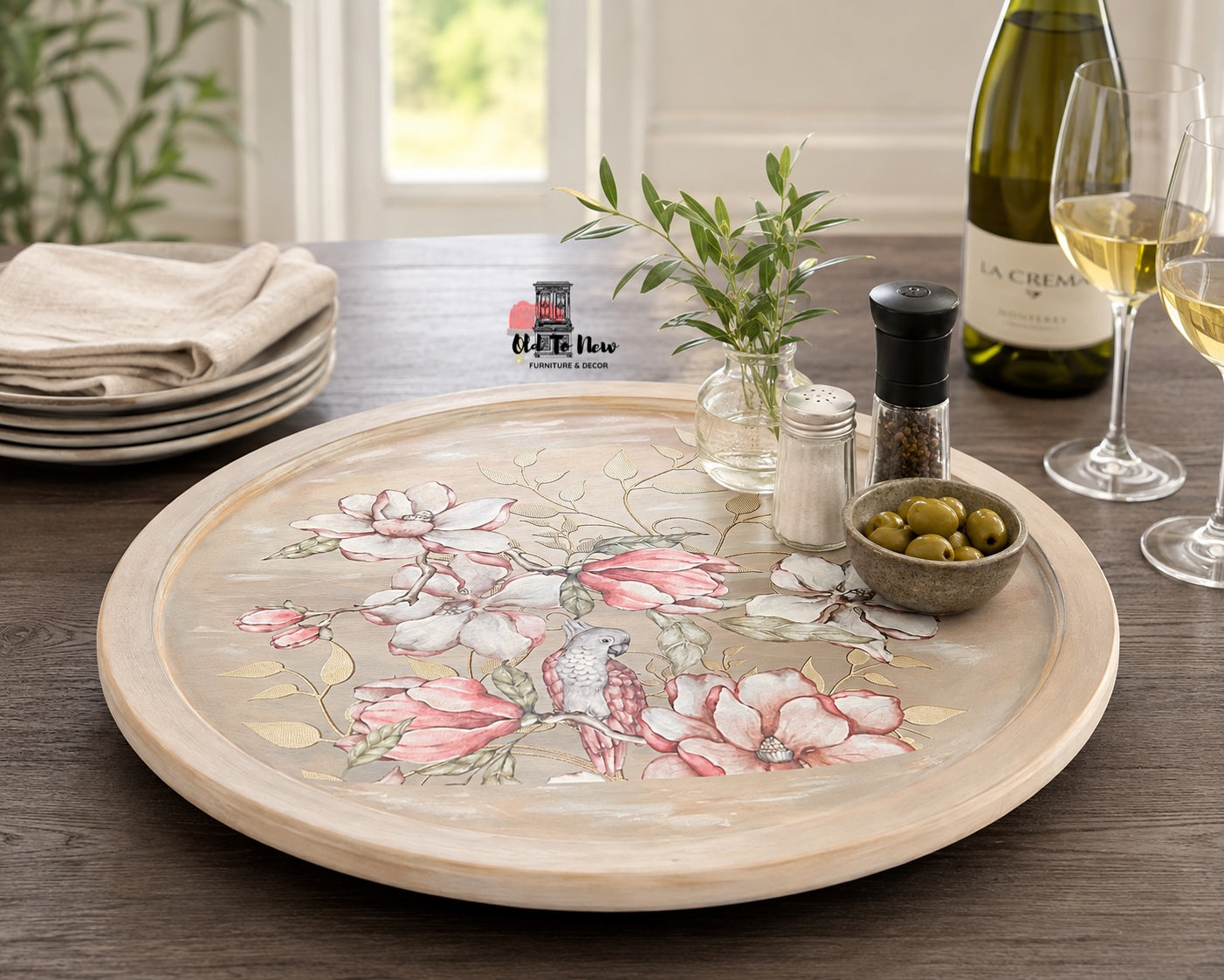 A round wooden serving tray with a floral and bird illustration, styled on a dark wood table with a wine bottle, glasses, olives, and a small vase of greenery.