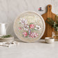 A round decorative serving platter with a distressed cream border and a floral design featuring pink, white, and green magnolia blossoms, styled on a marble countertop with a wooden cutting board, white pitcher, and small bowls.