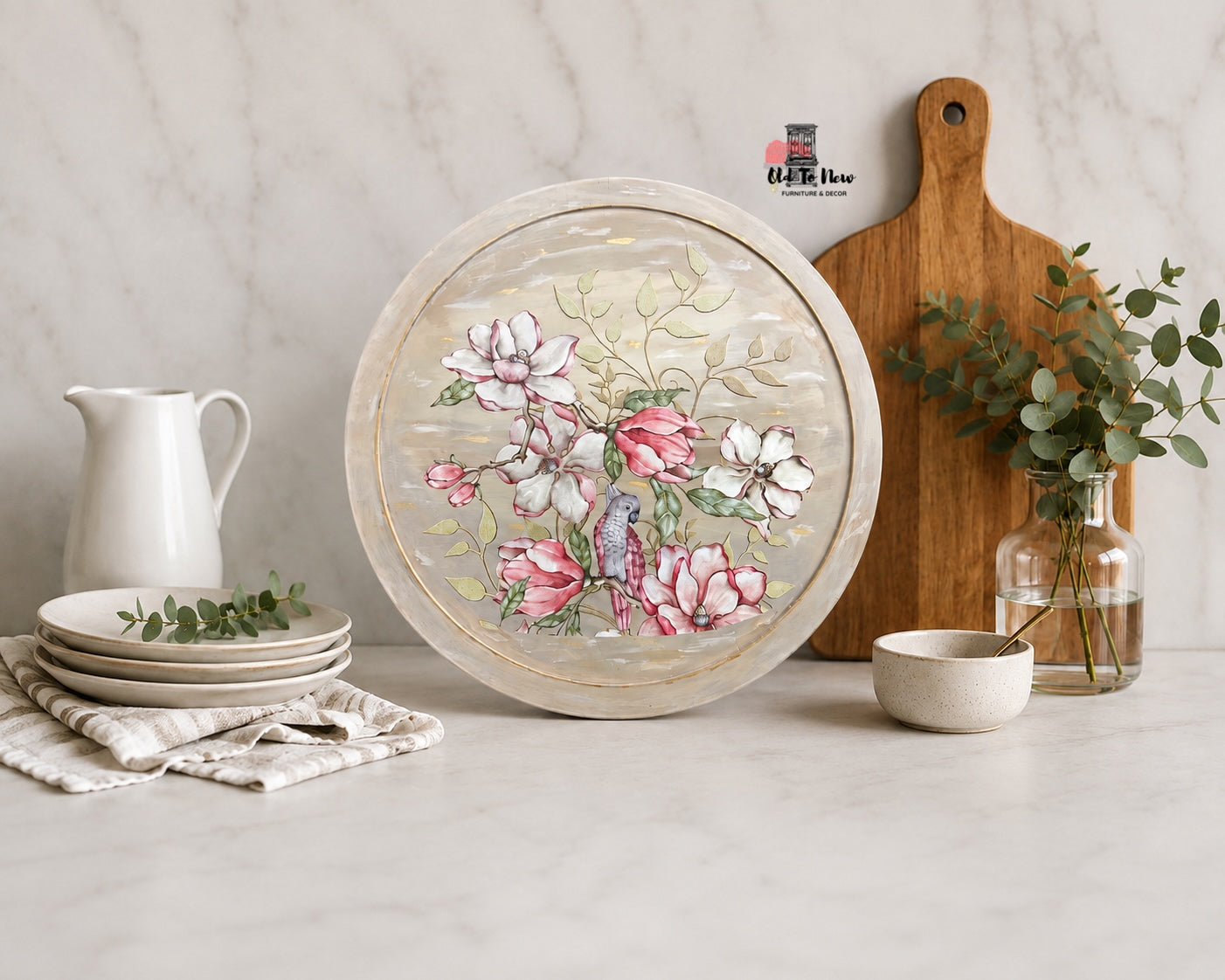 A round decorative serving platter with a distressed cream border and a floral design featuring pink, white, and green magnolia blossoms, styled on a marble countertop with a wooden cutting board, white pitcher, and small bowls.