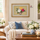 A framed floral needlepoint or embroidery artwork hanging on a cream wall above a white sofa in a styled living room. The artwork shows a bouquet of yellow, white, and pink flowers with green leaves in a brown vase on a beige background with a gold frame.