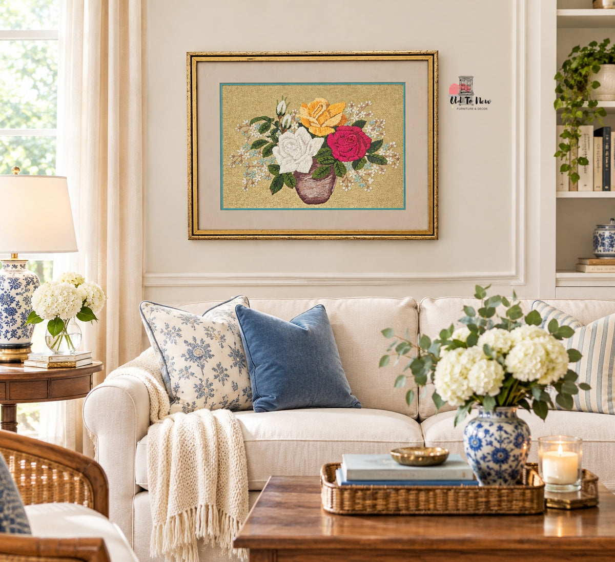 A framed floral needlepoint or embroidery artwork hanging on a cream wall above a white sofa in a styled living room. The artwork shows a bouquet of yellow, white, and pink flowers with green leaves in a brown vase on a beige background with a gold frame.