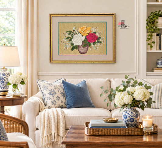 A framed floral needlepoint or embroidery artwork hanging on a cream wall above a white sofa in a styled living room. The artwork shows a bouquet of yellow, white, and pink flowers with green leaves in a brown vase on a beige background with a gold frame.
