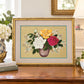 A framed floral needlepoint or embroidery artwork showing yellow, white, and pink roses in a vase, displayed on a wooden table in a styled home interior with books, flowers, and a lamp.