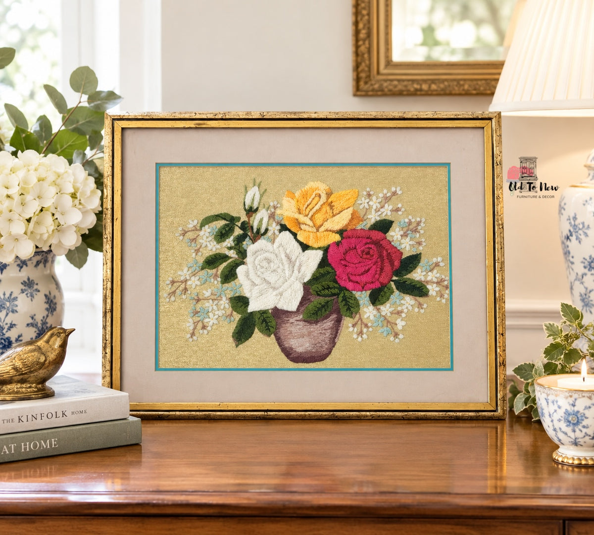 A framed floral needlepoint or embroidery artwork showing yellow, white, and pink roses in a vase, displayed on a wooden table in a styled home interior with books, flowers, and a lamp.