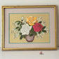 A framed needlepoint or embroidered floral artwork showing a bouquet of white, yellow, and pink roses with green leaves in a brown vase, set against a textured gold background. The piece is matted with a beige border and enclosed in a distressed gold frame, photographed against a plain light background.