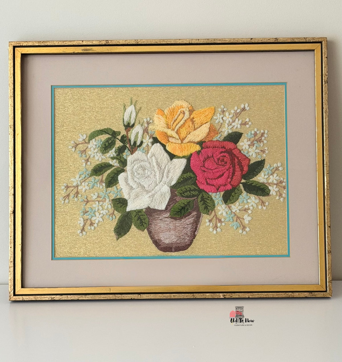 A framed needlepoint or embroidered floral artwork showing a bouquet of white, yellow, and pink roses with green leaves in a brown vase, set against a textured gold background. The piece is matted with a beige border and enclosed in a distressed gold frame, photographed against a plain light background.