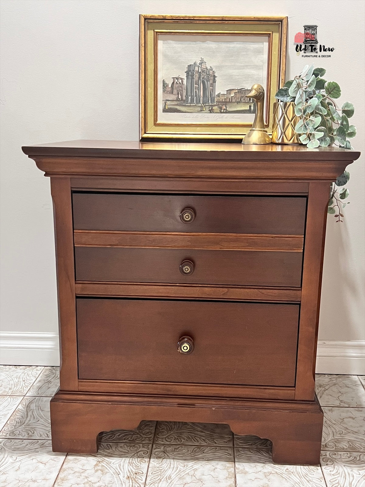 Wooden nightstand with three drawers, framed picture, and plant on top against a white wall. Old to New Furniture, Canada 