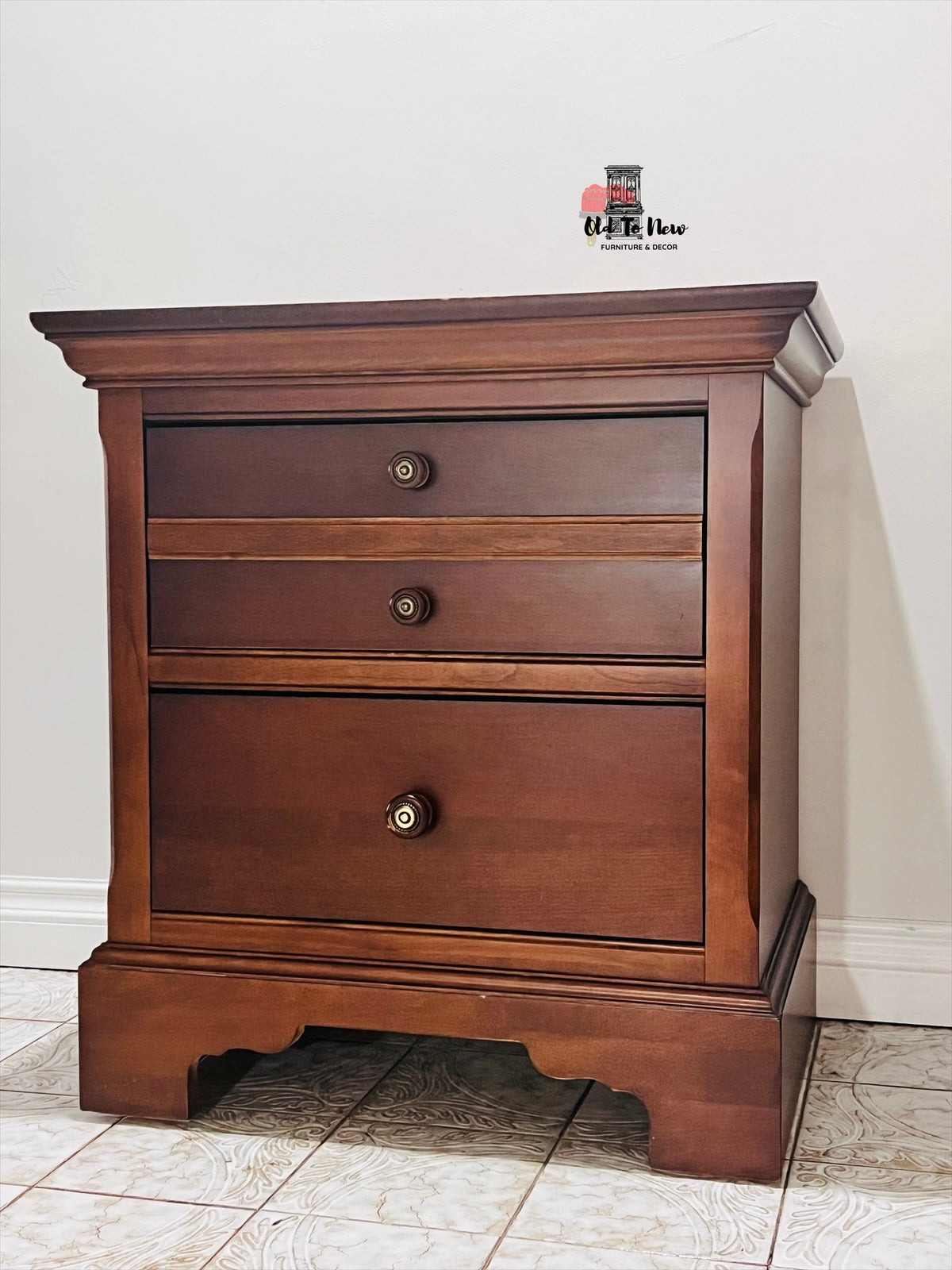 Wooden nightstand with three drawers on a tiled floor.
