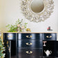 Black dresser with gold handles in a room with decorative mirror and plants.